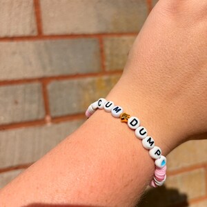 Op de afbeelding: Een kralenarmband met witte ronde kralen met zwarte letters die de tekst CUM DUMP vormen, afgewisseld met een kleine goudkleurige stervormige kraal en diverse roze en witte platte schijfvormige tussenkralen.