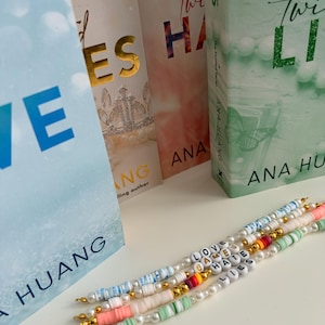 May include: Four beaded bracelets featuring white pearls, gold-tone spacers, and coloured clay discs. The bracelets spell out LOVE, GAMES, HATE, and LIES in white letter beads. They are positioned in front of four books by Ana Huang.