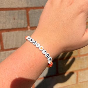 May include: A beaded bracelet featuring white circular beads with black letters spelling out the phrase ANAL TOY, interspersed with small orange heart-shaped beads and orange disc-shaped spacer beads on an elastic cord.