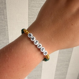 Könnte beinhalten: Ein Perlenarmband mit weißen runden Perlen, auf denen der schwarze Schriftzug MASTER aufgedruckt ist, durchsetzt mit metallisch goldenen kugelförmigen Perlen und dunkelgrünen scheibenförmigen Perlen in einem sich wiederholenden Muster.