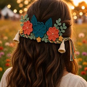 May include: A crochet hair accessory featuring a braided band adorned with two orange flowers, one teal flower, small yellow flowers, and green leaf sprigs. The piece includes two cream-colored bell-shaped tassels and dangling multi-colored yarn strands.