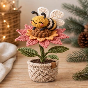 Op de afbeelding: Een gehaakte amigurumi-bij met een geel en zwart gestreept lichaam en witte vleugels, rustend op een grote roze en oranje bloem. De bloem heeft een groene steel met twee bladeren en staat in een kleine, getextureerde beige gehaakte pot.