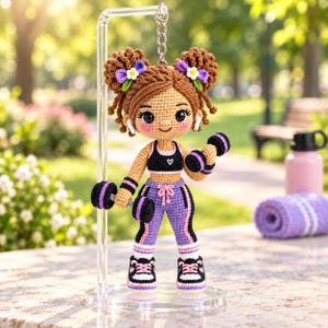 Pode incluir: Um porta-chaves de boneca em croché com uma personagem de cabelo castanho com dois ganchos, vestindo um top desportivo preto com um coração branco, leggings roxas com riscas pretas e sapatilhas pretas. A boneca segura dois pequenos halteres.