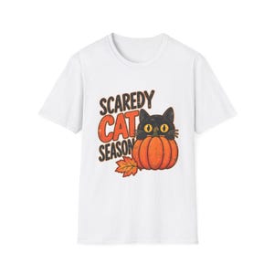 May include: A white short-sleeve t-shirt featuring a graphic of a black cat with large yellow eyes peeking out from behind an orange pumpkin. The text "SCAREDY CAT SEASON" is printed in black and orange, accompanied by a single orange maple leaf.