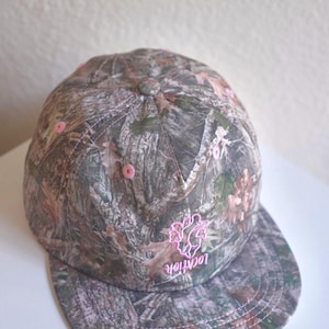 May include: A baseball cap featuring a forest camouflage pattern in shades of brown, green, and grey. The cap includes pink embroidered eyelets, a pink embroidered heart-shaped anatomical design, and the word "location" in pink embroidery.