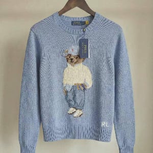 Può includere: Un maglione girocollo azzurro con un motivo lavorato a maglia raffigurante un orsacchiotto che indossa un berretto, un maglione color crema e pantaloni blu. L'orso è centrato sul petto. Il maglione ha collo, polsini e orlo a coste, con le lettere RL ricamate in bianco in basso a sinistra. È attaccata un'etichetta del marchio Polo Ralph Lauren.