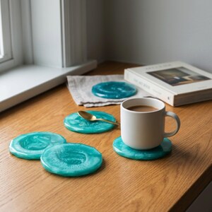 May include: A set of four round, teal-colored coasters with a swirling, marbled finish. One coaster supports a white ceramic mug filled with a light brown beverage, while a small gold-toned spoon rests on another coaster nearby.