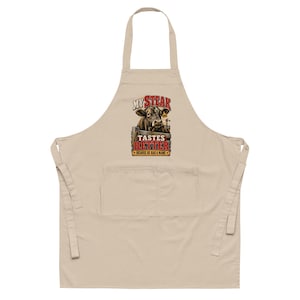 May include: A beige apron featuring a graphic of a cow behind a wooden fence with the text "MY STEAK TASTES BETTER BECAUSE HE HAD A NAME" in red and yellow lettering. The apron includes a neck loop, waist ties, and a large front pocket.
