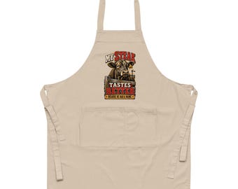 Op de afbeelding: Een beige schort met een afbeelding van een koe achter een houten hek met de tekst "MY STEAK TASTES BETTER BECAUSE HE HAD A NAME" in rode en gele letters. Het schort is voorzien van een nekband, taillebanden en een grote voorzak.