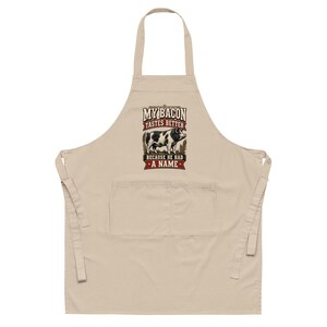 May include: A beige apron featuring a graphic of a black and white spotted pig in the center. Above the pig, the text reads MY BACON TASTES BETTER, and below the pig, the text reads BECAUSE HE HAD A NAME. The apron includes a large front pocket.