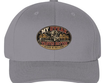 May include: A grey baseball cap featuring an oval graphic on the front. The graphic depicts a cow with an ear tag labelled "EARL" in a fenced area, with the text "MY STEAK TASTES BETTER BECAUSE HE HAD A NAME" printed in red and yellow lettering.