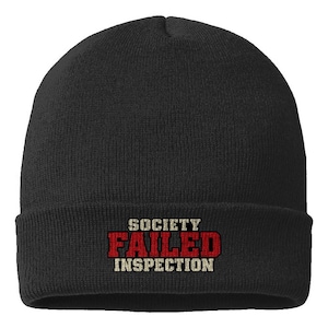 May include: A black knit beanie featuring the text "SOCIETY FAILED INSPECTION" embroidered on the front. The word "SOCIETY" and "INSPECTION" are in a light beige font, while the word "FAILED" is in a bold, red, block-style font.