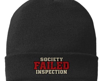 May include: A black knit beanie featuring the text "SOCIETY FAILED INSPECTION" embroidered on the front. The word "SOCIETY" and "INSPECTION" are in a light beige font, while the word "FAILED" is in a bold, red, block-style font.