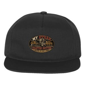 May include: A black baseball cap featuring a central oval graphic of a cow behind a wooden fence. The text reads MY STEAK TASTES BETTER BECAUSE HE HAD A NAME. The cow has an ear tag labeled EARL. The design uses brown, red, and yellow tones.