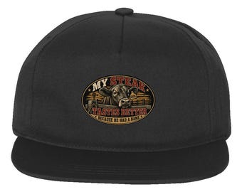 May include: A black baseball cap featuring a central oval graphic of a cow behind a wooden fence. The text reads MY STEAK TASTES BETTER BECAUSE HE HAD A NAME. The cow has an ear tag labeled EARL. The design uses brown, red, and yellow tones.