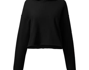 May include: A solid black cropped hoodie featuring long sleeves with ribbed cuffs, a hood with a drawstring-free neckline, and a raw-cut hemline. The garment has a plain front and a subtle graphic detail visible on the lower portion of the left sleeve.