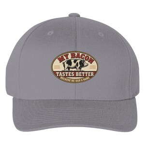 May include: A grey baseball cap featuring an oval patch on the front. The patch displays a black and white pig illustration with the text MY BACON TASTES BETTER BECAUSE HE HAD A NAME in red and black lettering against a cream background.