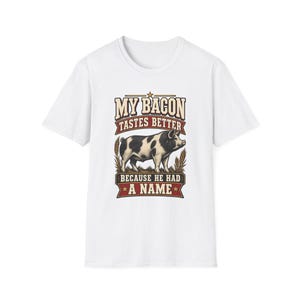 Könnte beinhalten: Ein weißes Kurzarm-T-Shirt mit einer Grafik eines schwarz-weiß gefleckten Schweins, das in Weizen steht. Der Text lautet MY BACON TASTES BETTER BECAUSE HE HAD A NAME in roter, weißer und brauner Schrift mit einem kleinen Stern über dem Schwein.