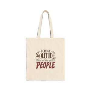 May include: A beige canvas tote bag featuring dark brown and red text that reads I CHOSE SOLITUDE because the other option was PEOPLE. The bag has two long shoulder straps and a rectangular body, presenting a clear and legible graphic design.