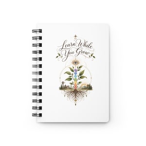 May include: A white spiral-bound notebook featuring a central graphic of a plant with seven colored chakra circles along its stem, roots at the base, and an eye symbol above. The text "Learn While You Grow" is printed in black cursive script.