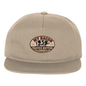 May include: A beige baseball cap featuring a central oval graphic with a black and white pig illustration. The text reads MY BACON TASTES BETTER BECAUSE HE HAD A NAME in red and black lettering against a cream background with red accents.