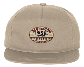 May include: A beige baseball cap featuring a central oval graphic with a black and white pig illustration. The text reads MY BACON TASTES BETTER BECAUSE HE HAD A NAME in red and black lettering against a cream background with red accents.