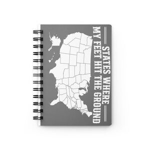 May include: A gray spiral-bound notebook featuring a white outline map of the United States on the front cover. The text printed in white capital letters reads: STATES WHERE MY FEET HIT THE GROUND. The notebook has a black metal wire binding.