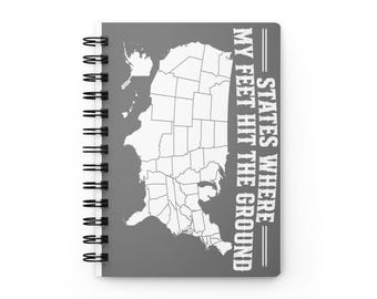 May include: A gray spiral-bound notebook featuring a white outline map of the United States on the front cover. The text printed in white capital letters reads: STATES WHERE MY FEET HIT THE GROUND. The notebook has a black metal wire binding.