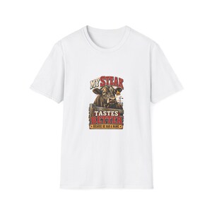 May include: A white short-sleeved t-shirt featuring a graphic of a brown cow behind a wooden fence. The text reads MY STEAK TASTES BETTER BECAUSE HE HAD A NAME. The design includes red and yellow lettering with a small windmill in the background.