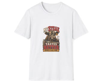May include: A white short-sleeved t-shirt featuring a graphic of a brown cow behind a wooden fence. The text reads MY STEAK TASTES BETTER BECAUSE HE HAD A NAME. The design includes red and yellow lettering with a small windmill in the background.