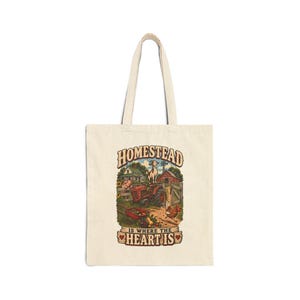 May include: A beige canvas tote bag featuring a colorful graphic of a farm scene with a red tractor, a goat, a chicken, and a house. The text reads HOMESTEAD IS WHERE THE HEART IS, with small red hearts flanking the bottom line of the printed design.