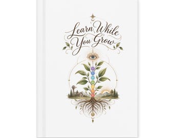 May include: A white hardcover journal featuring the text Learn While You Grow in elegant script. The design includes a central plant with seven colored chakra circles on its stem, roots extending downward, and a landscape with a house and sun.