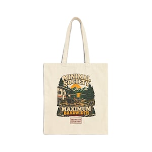 May include: A cream-colored canvas tote bag featuring a graphic illustration of a person sitting in a chair by a camper near a lake and mountains. The text reads MINIMAL SOCIETY MAXIMUM BANDWIDTH and UNCIVILIZED INTENTIONS in a rectangular box.