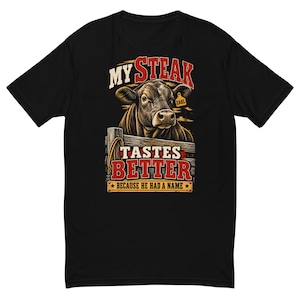 Könnte beinhalten: Ein schwarzes Kurzarm-T-Shirt mit einer Grafik einer Kuh hinter einem Holzzaun. Der Text lautet MY STEAK TASTES BETTER BECAUSE HE HAD A NAME. Die Kuh trägt eine Ohrmarke mit der Aufschrift EARL. Das Design verwendet rote, weiße und braune Farben.