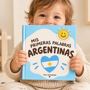 Puede incluir: Un libro de tapa dura cuadrado con una cubierta azul claro que presenta una forma de nube blanca, un sol amarillo sonriente y un corazón con la bandera argentina. El texto dice Mis Primeras Palabras Argentinas y Casa Mushaboom.