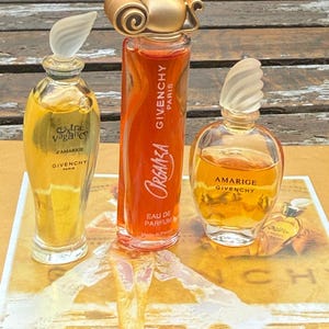 May include: Three glass perfume bottles by Givenchy. The left bottle is tall and slender with Extravagance d'Amarige text. The center bottle is tall with an orange liquid and Organza Givenchy Paris text. The right bottle is round with Amarige Givenchy text.