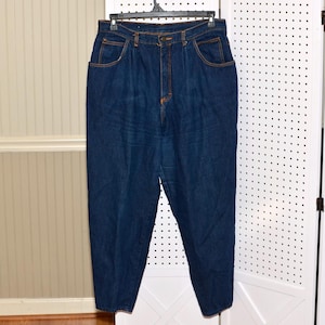 May include: High-waisted dark blue denim jeans featuring a pleated front design, a button and zipper fly closure, two side pockets, and contrasting orange stitching throughout the garment. The jeans have a tapered leg silhouette and a classic fit.