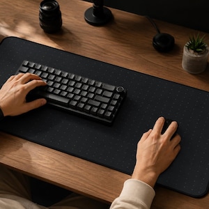May include: A large rectangular black desk mat with a subtle white dot grid pattern, featuring a compact black mechanical keyboard and a black computer mouse positioned on its surface for use in a workspace environment.