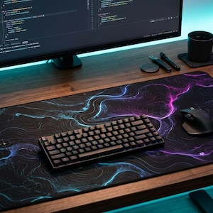 Puede incluir: Una alfombrilla de escritorio rectangular con fondo negro y líneas de contorno topográficas vibrantes en tonos azul neón y magenta. La alfombrilla está sobre una superficie de madera, sosteniendo un teclado mecánico negro y un ratón inalámbrico.