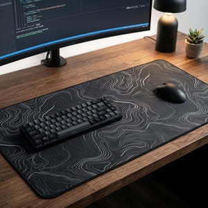 May include: A rectangular desk mat featuring a black surface with a white topographic map line pattern. The mat is positioned on a wooden desk surface, supporting a black mechanical keyboard and a black computer mouse for computer use.