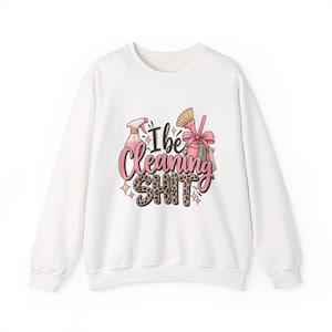 May include: A white long-sleeve sweatshirt featuring a graphic print with the text "I be Cleaning SHIT". The design includes illustrations of a pink spray bottle, a cleaning brush with a pink bow, and leopard print lettering for the word SHIT.