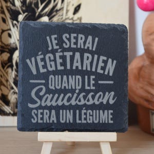 Peut inclure: Un dessous de verre carré en ardoise noire gravé du texte JE SERAI VÉGÉTARIEN QUAND LE SAUCISSON SERA UN LÉGUME dans un mélange de polices sans-serif et script. Le dessous de verre présente des bords naturels et repose sur un petit chevalet en bois.
