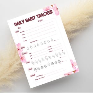 Op de afbeelding: Een wit dagelijks gewoonte-trackerblad met roze kersenbloesemillustraties in de hoeken. De tekst luidt DAILY HABIT TRACKER, Weight, Day, Food, Water, Exercise, Steps, Sleep en Overall, met iconen voor water, stappen en slaap.