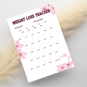 Puede incluir: Una hoja de papel blanca para el seguimiento de la pérdida de peso con texto en rojo oscuro que dice WEIGHT LOSS TRACKER, Starting Date y Starting Weight. El documento incluye una cuadrícula de casillas numeradas del 1 al 30, decorada con flores de cerezo rosas.