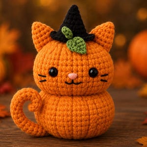 May include: A crocheted orange cat shaped like a pumpkin with a black witch hat and a small green leaf on top. The cat has black button eyes, a pink nose, and stitched whiskers. It features a curled tail and a round, textured body.