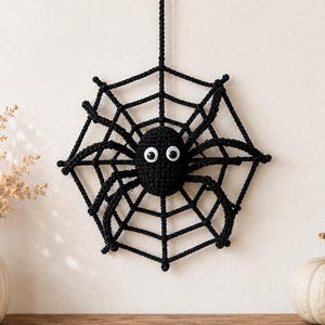 Peut inclure: Une araignée crochetée noire suspendue à une toile crochetée noire assortie. L'araignée a un corps rond avec huit pattes et deux grands yeux mobiles blancs avec des pupilles noires. L'ensemble est réalisé en fil de laine noir épais.