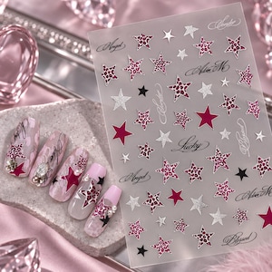 May include: A sheet of nail art stickers featuring various star shapes in solid magenta, silver glitter, and pink leopard print patterns, interspersed with cursive text including Angel, Lucky, and Blessed, alongside five decorated artificial nails.