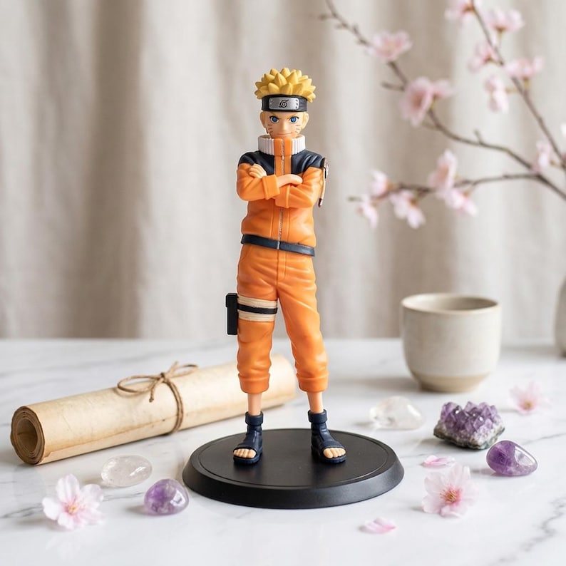 May include: A standing anime character figurine with spiky blonde hair, wearing a bright orange jumpsuit with dark blue accents and a headband featuring a metal plate with a spiral symbol. The figure stands on a circular black base.
