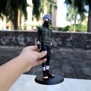 May include: A standing anime character figurine featuring spiky silver hair, a black face mask, and a headband. The figure wears a dark blue long-sleeved shirt, a green tactical vest with multiple pockets, dark pants, black gloves, and black boots.