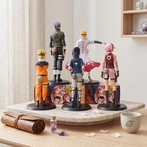 May include: Five anime character figurines standing on a circular display base. The figures include characters in orange, green, blue, white, and maroon outfits, each on a black circular stand. A brown scroll, a small glass vial, and a cup are nearby.